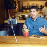 Social Media Influencer Or Food Blogger Creating Content Inside Small Restaurant - Man Sharing Online Food Review Using Smartphone On Tripod And Lavalier - Smiling Content Creator Vlogger Filming Video