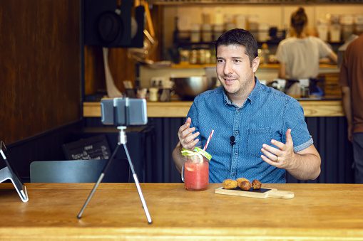 Social media influencer or food blogger creating content inside small restaurant - man sharing online food review using smartphone on tripod and lavalier - smiling content creator vlogger filming video